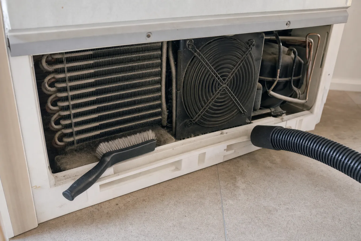 Kickplate removed from a white cabinet: dusty condenser coil, fan behind its wire guard and the compressor, a stiff brush and vacuum hose