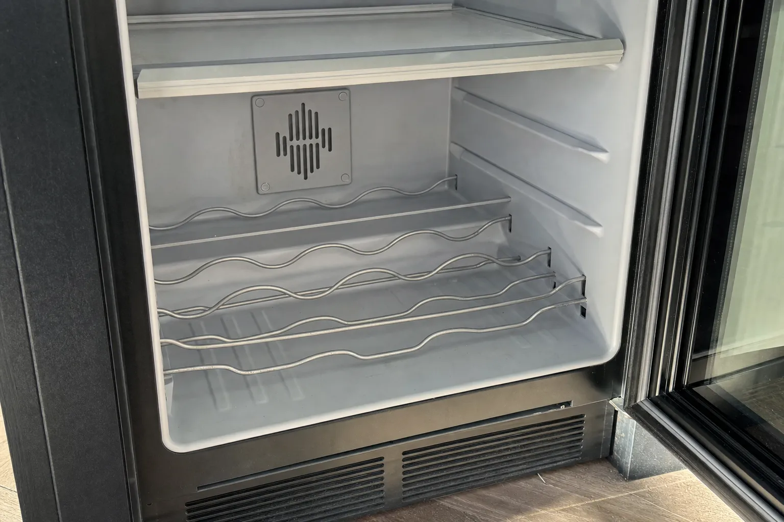 Dark undercounter unit with its glass door open: a glass shelf above four wavy wire bottle racks, a vent plate on the back wall